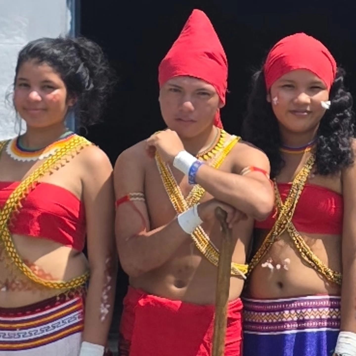 Tres jóvenes indígenas vistiendo trajes ceremoniales rojos con bordados tradicionales posan junto a una vivienda comunitaria
