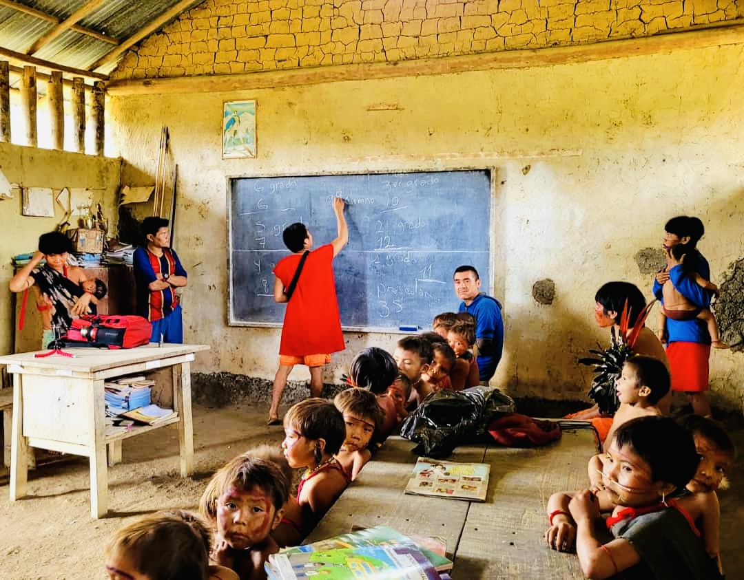 Aula de escuela comunitaria indígena con niñas y niños yanomami trabajando junto a un maestro frente a una pizarra