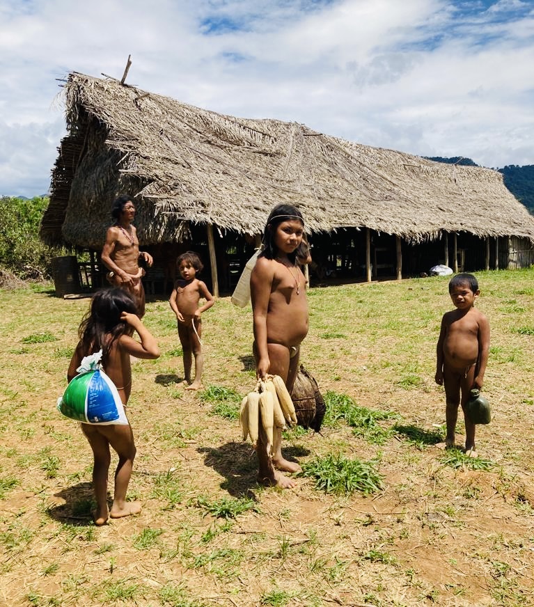 Niñas, niños y adulto del pueblo Hoti en su comunidad de la selva, frente a una vivienda tradicional de palma, cargando maíz y agua