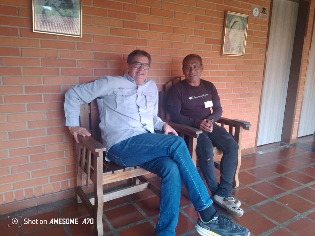 Vladimir Aguilar Castro sonriente, sentado en un banco de madera junto a un compañero indígena